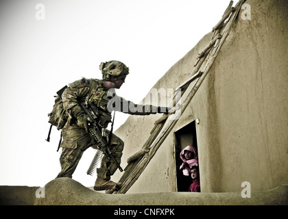 A US soldier climbs a ladder to set up a defensive watch position during an air assault mission while children watch through a window March 21, 2012 in Ghazni province, Afghanistan. The soldiers, alongside the Afghan National Army, searched two villages for insurgent activity and weapons caches. Stock Photo