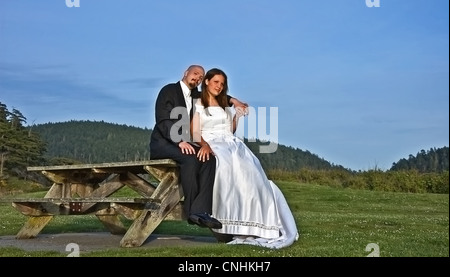 This young Caucasian bride and groom are sitting outdoors on a rustic picnic table with the groom's arm around the bride. Stock Photo