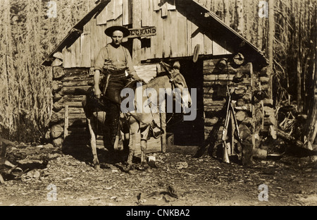 Heavily Armed Cowboy Riding Donkey at Log Cabin Stock Photo