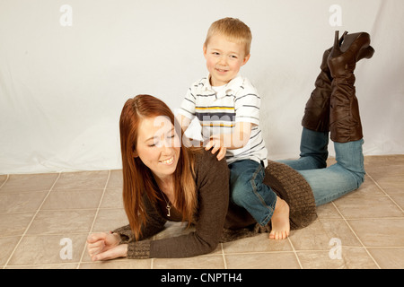 Pretty redheaded mother with her blond son sitting on her back while she lays down on the floor Stock Photo