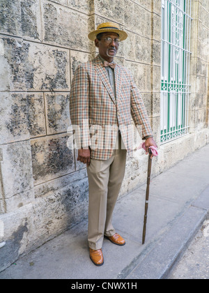 A dark elderly Cuban gentleman in a straw boater hat, checked suit and bow tie stands by a stone wall in Havana. Stock Photo