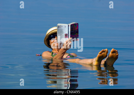 older woman wearing a straw hat and sunglasses is reading a book while floating in the Dead Sea on her back, Jordan Stock Photo