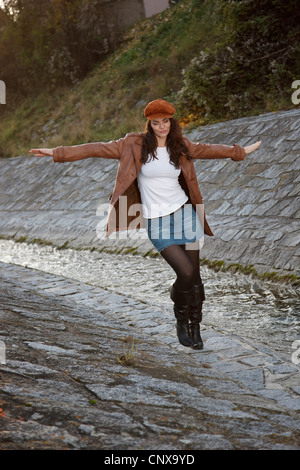 brunette young woman walking with the arms stretched out posing at the edge of a canalised brook in a mini skirt and leather boots Stock Photo