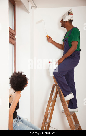A man standing on a ladder using a paint roller while a woman watches Stock Photo