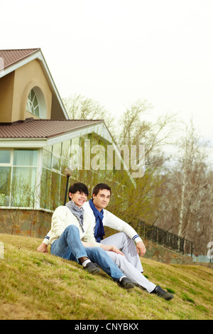 Tilt up of stylish homosexual lovers sitting on the grass in front of the townhouse Stock Photo