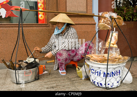 Horizontal portrait of a Saigonese street vendor making hot waffles from her yoke to sell sitting on the pavement in Ho Chi Minh, Vietnam. Stock Photo