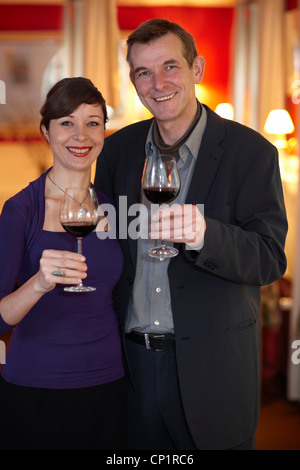 Happy smiling middle aged couple standing celebrating with glasses of red wine in plush red interior. Stock Photo