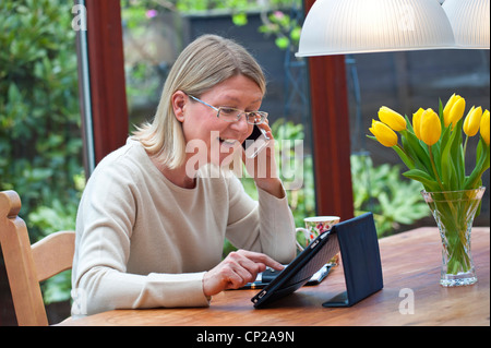 Blond woman relaxing at home in garden conservatory using her iPad tablet computer and talking on her iPhone 4s smartphone Stock Photo