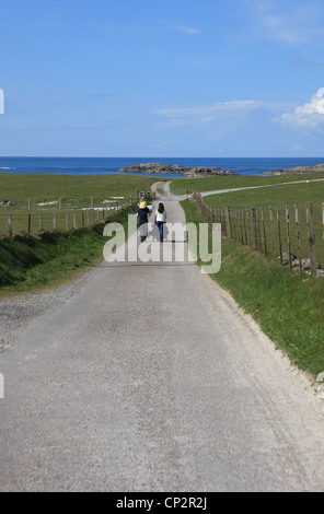 Family walking down a country road towards the sea. There is a father with daughter on his shoulders and a mother pushing a pram Stock Photo