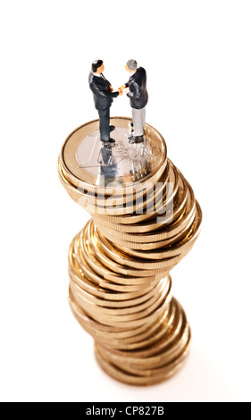 Two business men standing on a wobbly pile of € coins and shake hands. Stock Photo