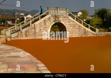 Man walking over stone bridge with bright red algae bloom scum on Longxi river in Chengkan village Huangshan Peoples Republic of China Stock Photo