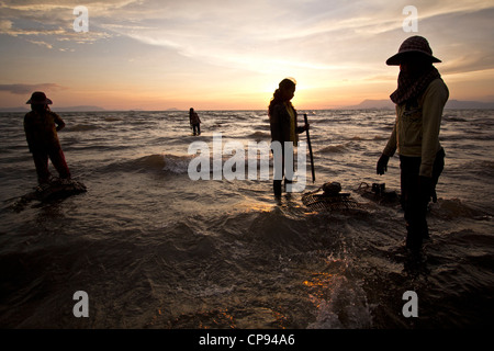 Female crab sellers stand in the water in Kep, Cambodia, with the sun setting behind them over the Gulf of Thailand Stock Photo
