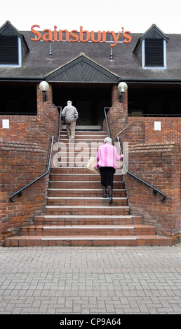 Elderly lady holding shopping bag climbing steps to a Sainsburys supermarket holding handrail Stock Photo