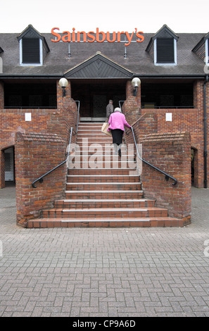 Elderly lady holding shopping bag climbing steps to a Sainsburys supermarket holding handrail Stock Photo