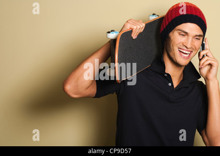 Portrait of good looking young caucasian man in black collar shirt wearing beanie using mobile phone and holding skateboard. Stock Photo