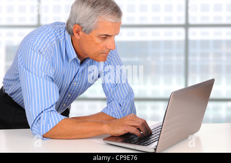 Smiling Middle Aged Businessman leaning on desk using laptop computer with large window background Stock Photo