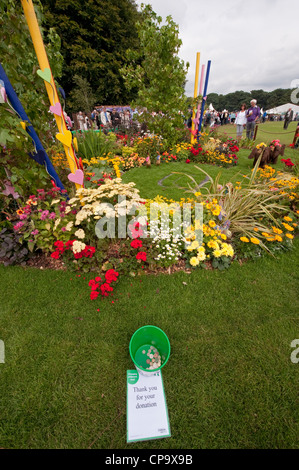 Bucket of money donations & visitors by beautiful colourful flowers in NSPCC's 'Hear our Cry' show garden - RHS Flower Show, Tatton Park, England, UK. Stock Photo