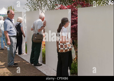 People peering in & viewing 'The Kaleidoscope' garden through gap in boundary wall (white panels) - RHS Flower Show, Tatton Park, Cheshire, England. Stock Photo