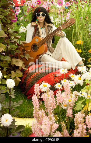 1960's flower power figure wearing shades, holding guitar, sitting on cushion by bright colourful flowers - RHS Flower Show, Tatton Park, Cheshire, UK Stock Photo