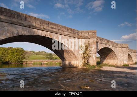 Sunny, scenic, rural riverside view of historic, stone, arched bridge spanning flowing water of River Wharfe - Burnsall, Yorkshire Dales, England, UK. Stock Photo