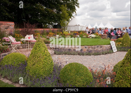 Visitors view beautiful flowers, lawn & small quiet patio seating area in  'Serenity' show garden - RHS Flower Show, Tatton Park, Cheshire, England. Stock Photo