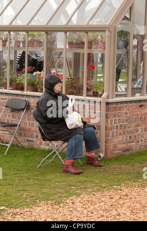 Sitting in rain (waterproof coat hood up) lady is wet & miserable on day out (by trade stand greenhouse) - RHS Tatton Park flower show, Cheshire, UK. Stock Photo