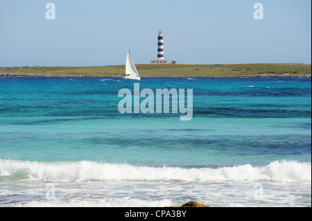 Cruising yacht sailing in turquoise sea past the striped lighthouse on Illa de l'Aire. Seen from the beach at Punta Prima, Menorca, Spain Stock Photo