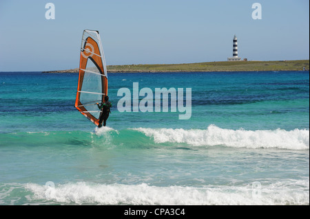 Windsurfer sailing in turquoise sea past the lighthouse on Isla del Aire, Punta Prima, Menorca, Spain Stock Photo