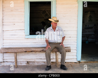 A 60-70 year old Cuban Hispanic male sits on a crude wooden bench on his front porch wearing a straw hat in Viñales, Cuba. Stock Photo