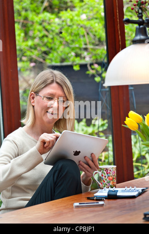 Blond woman relaxing at home in contemporary garden conservatory using her tablet iPad computer Stock Photo