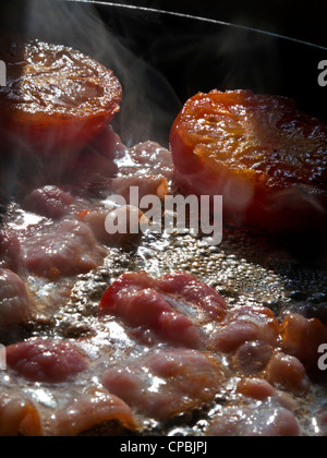 Shaft of sunlight illuminates streaky bacon rashers and tomatoes frying in hot pan Stock Photo