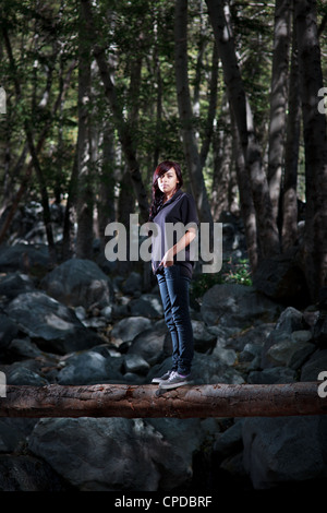 A young multi-ethnic woman with long dark hair stands on a log in a dark forest. Stock Photo