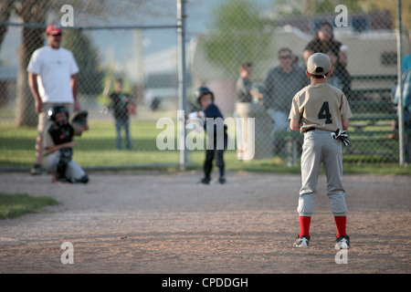 Little league baseball team players boy hits ball and runs to first base. Two teams in uniform. Small farming rural community. Stock Photo