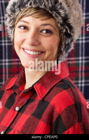 Wonderful smiling young woman wearing red and black checked lumberjack shirt and fur hat. Studio shot as close up portrait. Stock Photo