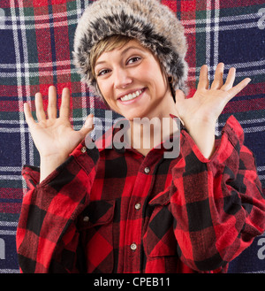 Smiling young woman wearing checkered lumberjack shirt and fur hat waving with both hands at the camera. Funny studio shot Stock Photo