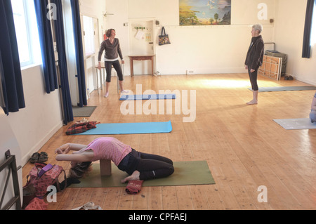 A woman in her mid forties warms up before a yoga class held in a village hall in Cornwall, UK. Other people just arriving Stock Photo