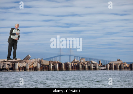 Oversized businessman standing on breakwater, holding briefcase stuffed with money Stock Photo