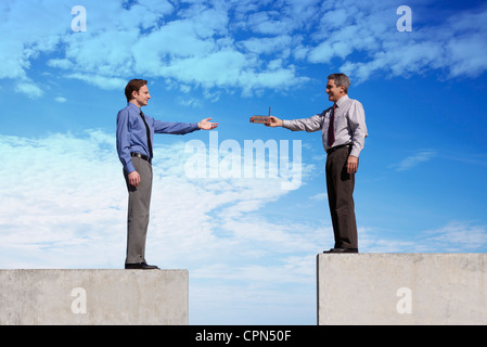 Businessmen standing on platforms, one handing the other a tiny factory Stock Photo