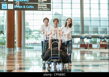 Brother and sister pushing younger sister on luggage cart in airport Stock Photo