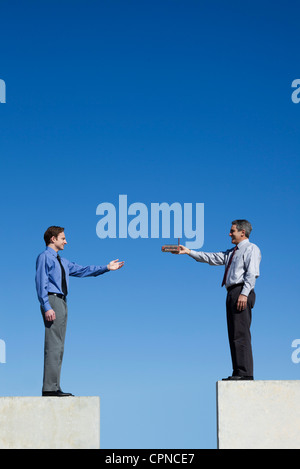 Businessmen standing on platforms, one handing the other a tiny factory Stock Photo