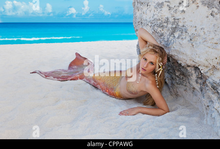 Young blond mermaid laying in the shade of a large rock on the beach in Cancun Mexico. Stock Photo