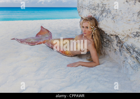 Young blond mermaid laying in the shade of a large rock on the beach in Cancun Mexico. Stock Photo
