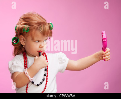 Little girl playing with mothers makeup and holding mirror on pink background Stock Photo