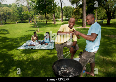 Two young African men barbeque together in a park with their friends sitting in the background Stock Photo