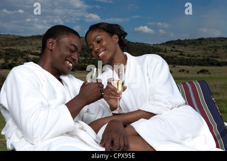 An African couple sit together outdoors wearing robes and holding champagne glasses Stock Photo