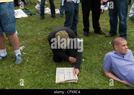 Man wearing flat cap on the ground reading the sporting pages of a newspaper, he is checking the runners and their odds. He has Swallow tattoo on his hand  he is a former sailor.  They are On The Hill, watching the horse racing Derby Day, Epsom Downs Surrey UK 2012  2010s  HOMER SYKES Stock Photo