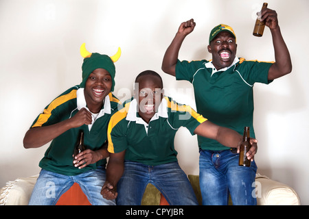 Three friends dressed in rugby uniforms watch a rugby game on TV whilst drinking beer Stock Photo