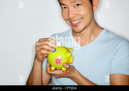 Portrait of asian man wearing light blue tee shirt holding a piggy bank and coin looking at camera smiling Stock Photo