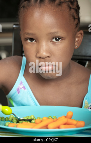 Young African child frowning at camera with a plate of vegetables in front of her Stock Photo
