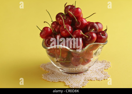 A scalloped clear glass bowl full of red cherries on a crocheted doily in a yellow background, vertical format. Stock Photo